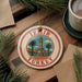 A circular ornament with the text "IZmir Turkey" and an illustration of a tower surrounded by palm trees, placed on a wooden surface with pine branches and a cup of coffee nearby.