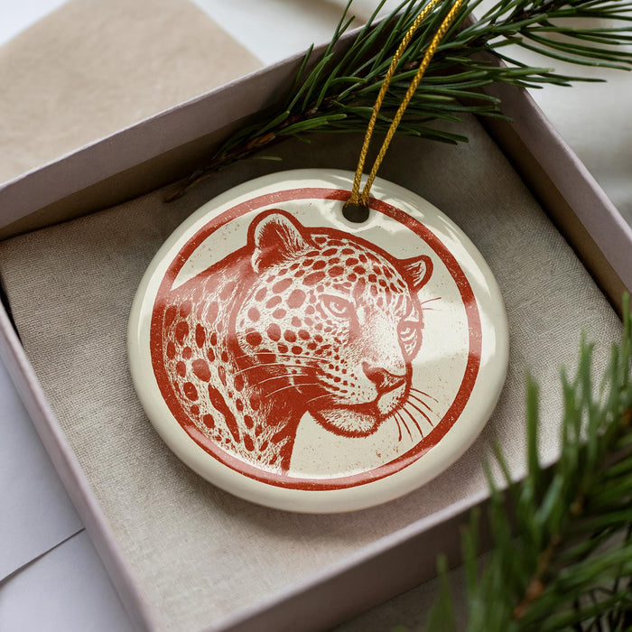 A ceramic ornament in the shape of a leopard's head, with a red and white color scheme, is displayed in a box surrounded by pine branches.