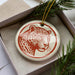 A ceramic ornament in the shape of a leopard's head, with a red and white color scheme, is displayed in a box surrounded by pine branches.