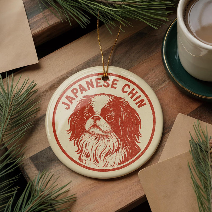 A circular ornament featuring a Japanese Chin dog is displayed on a wooden surface, accompanied by a cup of coffee and some pine branches.