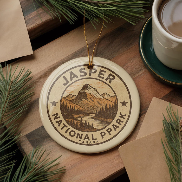 A circular ornament with the text "JASPER NATIONAL PARK" and a mountain landscape design hangs on a wooden surface, surrounded by pine branches and a cup of coffee.
