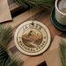 A circular ornament with the text "JASPER NATIONAL PARK" and a mountain landscape design hangs on a wooden surface, surrounded by pine branches and a cup of coffee.