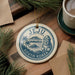 A decorative ornament featuring the text "JEJU SOUTH KOREA" and a stylized illustration of a mountain, palm tree, and sea, hanging on a wooden surface with pine branches and a cup of coffee nearby.