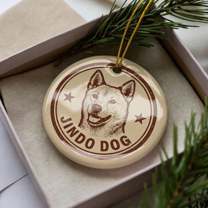A round ornament with a dog's face and the text "JINDO DOG" printed on it, placed inside a box with pine branches.