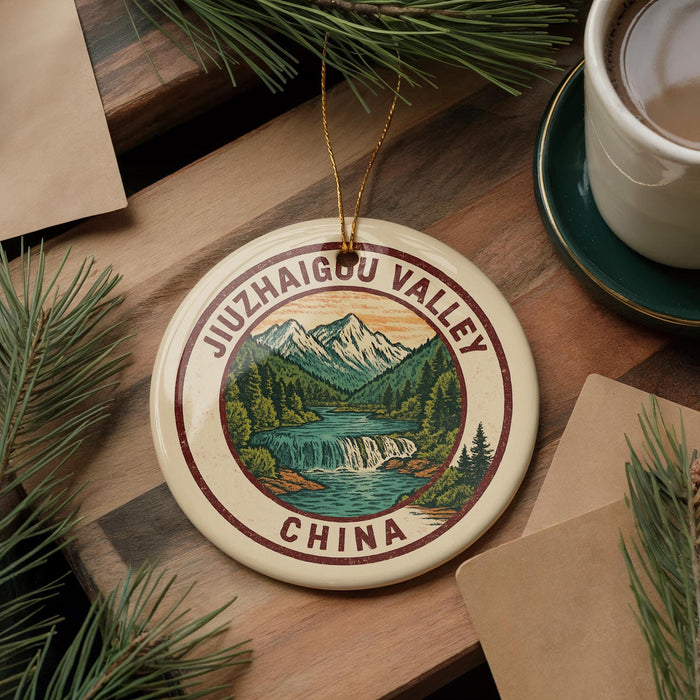A decorative ornament featuring a scenic mountain landscape with a waterfall, surrounded by pine trees, is displayed on a wooden surface alongside a cup of coffee and some papers.
