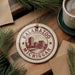 A circular ornament with the text "KALAMAZOO MICHIGAN" printed on it, surrounded by pine branches and a cup of coffee on a wooden surface.