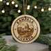 A circular ornament with the text "KAMPALA UGANDA" and an illustration of a cityscape is displayed on a table, surrounded by pine branches and twinkling lights.