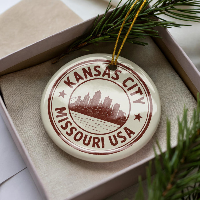 A circular ornament with the text "KANSAS CITY MISSOURI USA" printed on it, placed inside a box with pine branches.