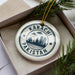 A circular ornament with the text "KARACHI PAKISTAN" printed on it, surrounded by a decorative border. The ornament is placed inside a box, with a sprig of greenery visible to the right.