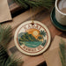A circular ornament with the text "Kauai Hawaii Us" and a tropical landscape design, including a mountain, palm tree, and ocean, is displayed on a wooden surface alongside a cup of coffee and some pine branches.