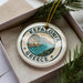 A decorative ornament in a box, featuring a circular design with the text "KEFALONIA GREECE" and an illustration of a coastal town.