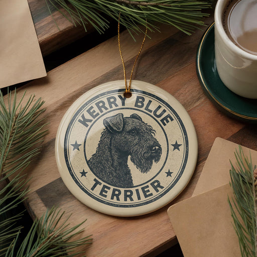 A ceramic ornament featuring a black and white illustration of a terrier dog, with the text "Kerry Blue Terrier" around the edge. The ornament is hanging on a wooden surface, with pine branches and a cup of coffee visible in the background.