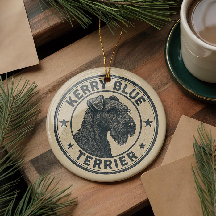 A ceramic ornament featuring a black and white illustration of a terrier dog, with the text "Kerry Blue Terrier" around the edge. The ornament is hanging on a wooden surface, with pine branches and a cup of coffee visible in the background.