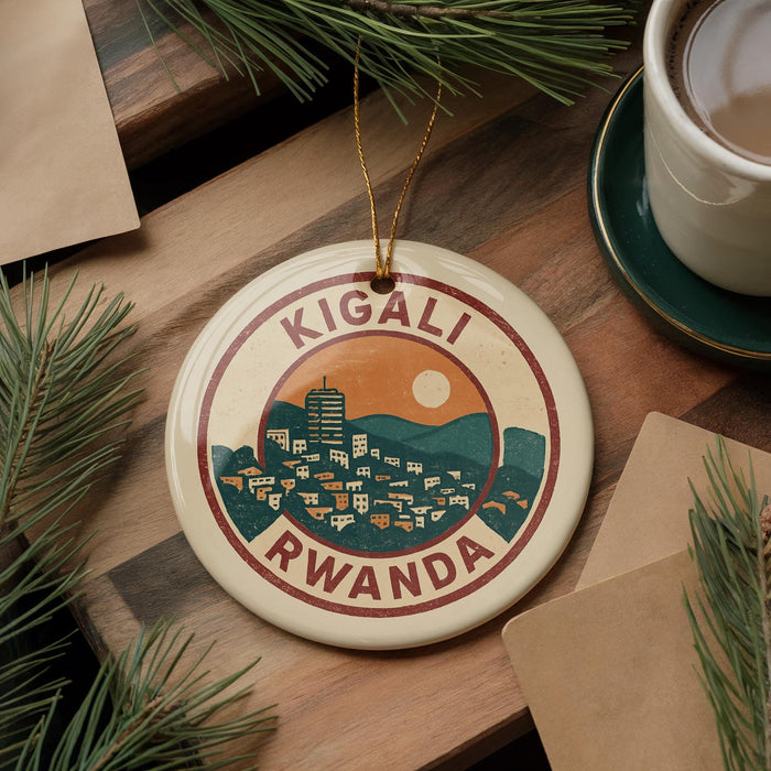 A decorative ornament featuring a circular design with a cityscape and the text "KIGALI RWANDA" is displayed on a wooden surface, surrounded by pine branches and a cup of coffee.