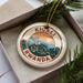 A decorative ornament with the text "KIGALI RWANDA" on it, placed in a box with pine branches.