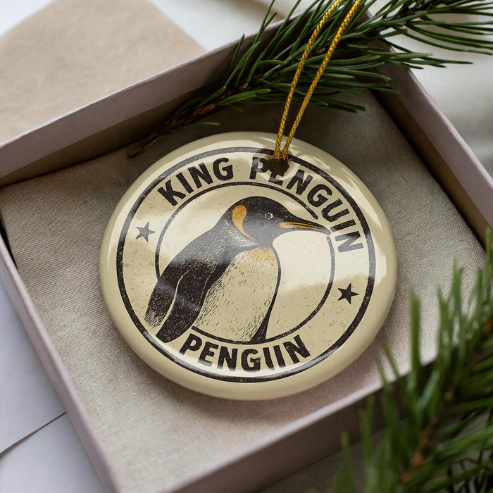 A round ornament with a penguin image and the text "KING PENGUIN PENGUIN" printed on it, placed inside a box with pine branches.