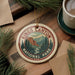 A circular ornament with the text "Kings Canyon National Park" and a mountain landscape design, placed on a wooden surface alongside pine branches and a cup of coffee.