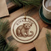 A circular ornament featuring a kinkajou monkey, a type of primate, is displayed on a wooden surface alongside a cup of coffee and some pine branches.