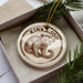 A circular ornament with a brown and white design featuring a small animal, likely a porcupine, is displayed in a box with pine branches.