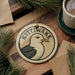 A circular ornament with a bird design and the word "KITTIWAKES" written on it, placed on a wooden surface next to a cup of coffee and pine branches.
