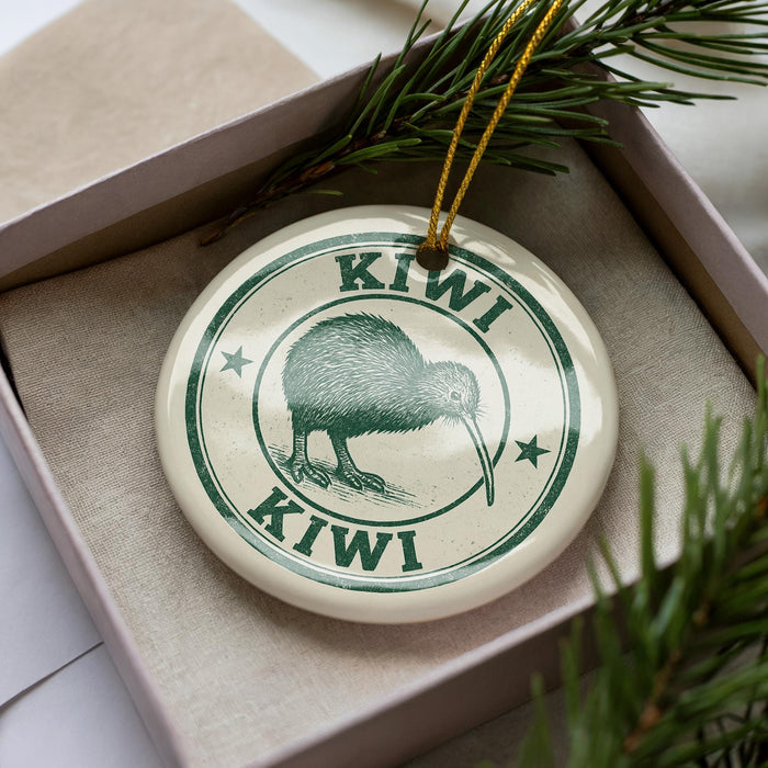 A circular ornament with the word "Kiwi" printed on it, featuring a kiwi bird illustration. The ornament is placed inside a box, surrounded by pine branches.