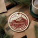 A circular ornament featuring a red and white illustration of the Kiyomizu-Dera temple in Kyoto, Japan, with the text "Kiyomizu-Dera" around the edge. The ornament is hanging from a string, and there is a cup of coffee and some pine branches in the background.