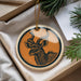 a decorative ornament with a detailed illustration of a horned animal, possibly a deer or a gazelle, on a circular base. The ornament is placed on a surface with some greenery, such as pine branches, and there is a white card or paper visible in the background.