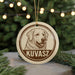 A circular ornament featuring a dog's head and the text "KUVASZ" in the center, hanging on a string against a blurred background of Christmas lights.