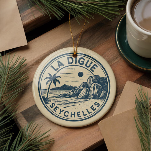A circular ornament with the text "LA DIGUE SEYCHELLES" and a palm tree illustration, placed on a wooden surface next to a cup of coffee and some pine branches.