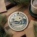 A circular ornament with the text "LA DIGUE SEYCHELLES" and a palm tree illustration, placed on a wooden surface next to a cup of coffee and some pine branches.