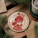 A red and white ornament featuring Ladybug, a character from the anime series "Ladybug", is displayed on a wooden surface alongside a cup of coffee and some pine branches.
