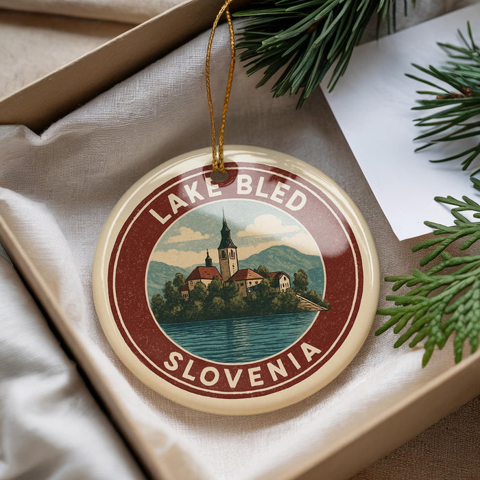 A round ornament with a picture of Lake Bled, Slovenia, is displayed in a box with pine branches.