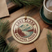 A circular ornament with the text "LAKELAND NATIONAL PARK USA" and a scenic illustration of Lake Clark National Park, surrounded by pine branches and a cup of coffee.
