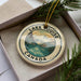 A circular ornament with the text "LAKELOUISE CANADA" and an illustration of a mountain range, placed in a box with pine branches.
