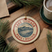 A circular ornament with the text "LAKE TITICACA PERU BOLIVIA" on it, surrounded by pine branches and a cup of coffee on a wooden surface.