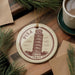 A circular ornament featuring the Leaning Tower of Pisa, a famous landmark in Italy, is displayed on a wooden surface alongside a cup of coffee and some pine branches.