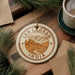 A circular ornament with the text "LEH LADAKH INDIA" printed on it, surrounded by pine branches and a cup of coffee on a wooden surface.