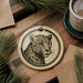 A ceramic ornament featuring a leopard's face is displayed on a wooden surface, surrounded by pine branches and a cup of coffee.