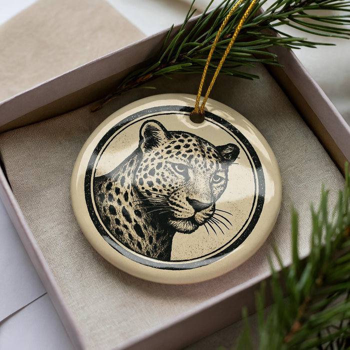 A ceramic ornament in the shape of a leopard's face, with a black and white design, is displayed in a box surrounded by pine branches.