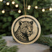 A round ornament featuring a leopard's face is displayed in front of a blurred background of Christmas lights.