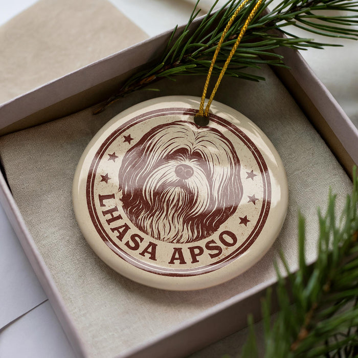 A circular ornament with a brown and white design featuring a dog's head and the text "LHASA APSPO" is displayed in a box with pine branches.