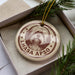 A circular ornament with a brown and white design featuring a dog's head and the text "LHASA APSPO" is displayed in a box with pine branches.