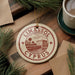 A circular ornament with the text "LIMASSOL" and "CYPRUS" printed on it, surrounded by pine branches and a cup of coffee on a wooden surface.