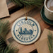 A circular ornament with the text "LINCOLN NEBDAKA USA" and a graphic of a city skyline is displayed on a wooden surface, surrounded by pine branches and a cup of coffee.