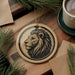 a circular ornament with a lion's head design, placed on a wooden surface alongside a cup of coffee and some pine branches.