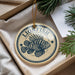A circular ornament with a lionfish design, hanging from a string, placed inside a box with pine branches.