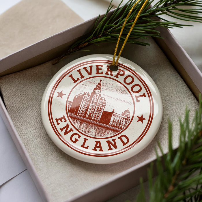 A circular ornament with the text "LIVERPOOL ENGLAND" printed on it, surrounded by a decorative border. The ornament is placed inside a box, with a sprig of greenery visible to the right.