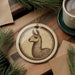 a round ornament with a brown and white illustration of a llama on a wooden surface, accompanied by a cup of coffee and some pine branches.