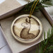 A ceramic ornament featuring a brown llama is displayed in a box, surrounded by pine branches.