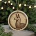 A round ornament featuring a brown llama illustration is displayed in front of a blurred background of Christmas lights.